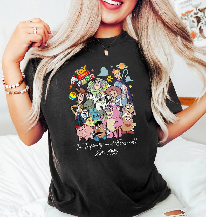May include: Black t-shirt featuring a colorful graphic of Toy Story characters, including Woody, Buzz Lightyear, and others. The shirt has the text "Toy Story Land" at the top and "To Infinity and Beyond! Est 1995" below the characters.