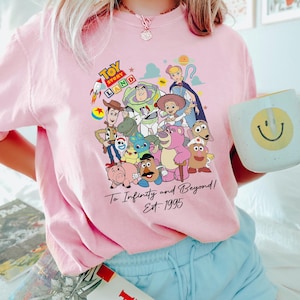 May include: Pink t-shirt featuring a colorful graphic of Toy Story characters, including Woody, Buzz Lightyear, and others. The text "Toy Story Land" and "To Infinity and Beyond! Est. 1995" are also visible. A fun, nostalgic design.