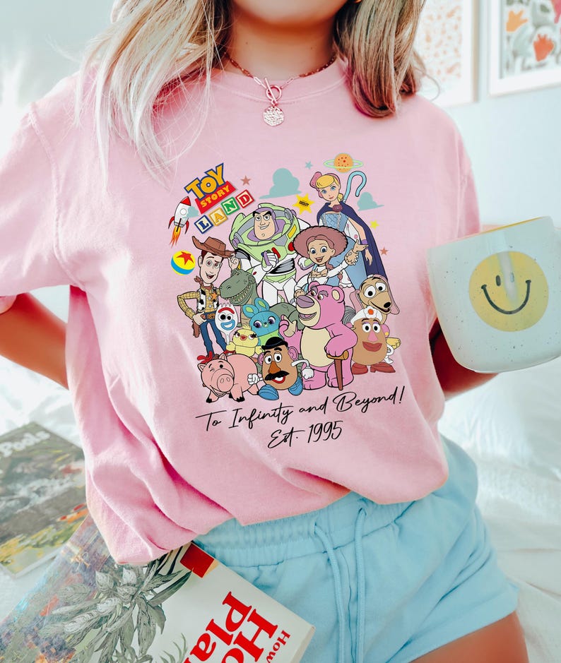 May include: Pink t-shirt featuring a colorful graphic of Toy Story characters, including Woody, Buzz Lightyear, and others. The text "Toy Story Land" and "To Infinity and Beyond! Est. 1995" are also visible. A fun, nostalgic design.