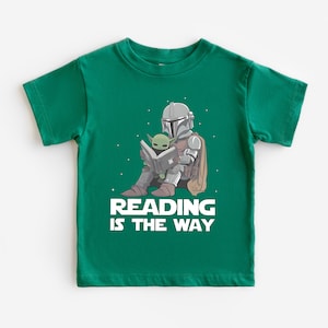 May include: A green t-shirt featuring a graphic of The Mandalorian and Grogu reading a book. The Mandalorian is in his armor, and Grogu is sitting on his lap. The text "READING IS THE WAY" is printed in white below the image.