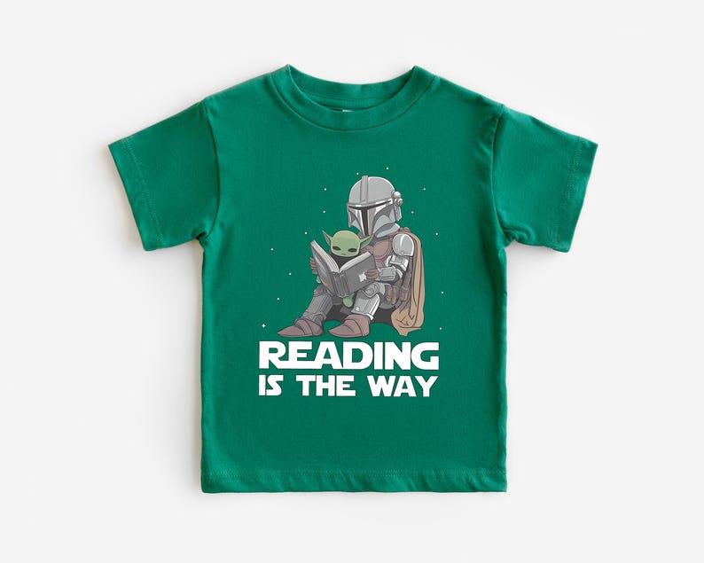 May include: A green t-shirt featuring a graphic of The Mandalorian and Grogu reading a book. The Mandalorian is in his armor, and Grogu is sitting on his lap. The text "READING IS THE WAY" is printed in white below the image.