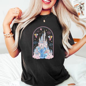 K&ouml;nnte beinhalten: Schwarzes T-Shirt mit einem farbenfrohen Aufdruck eines Schlosses, Feuerwerk und einer blauen Cartoon-Figur. Das Design ist von einem rosa Bogen mit floralen Akzenten umrahmt. Das T-Shirt hat einen l&auml;ssigen Stil.