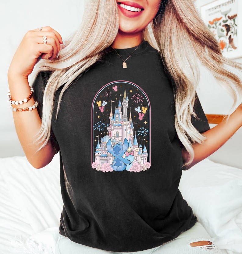 K&ouml;nnte beinhalten: Schwarzes T-Shirt mit einem farbenfrohen Aufdruck eines Schlosses, Feuerwerk und einer blauen Cartoon-Figur. Das Design ist von einem rosa Bogen mit floralen Akzenten umrahmt. Das T-Shirt hat einen l&auml;ssigen Stil.