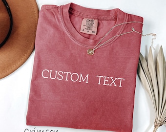 May include: Crimson-colored t-shirt with the words "CUSTOM TEXT" printed in white. A gold necklace with a pendant rests on the shirt. A brown hat and dried leaves are in the background.