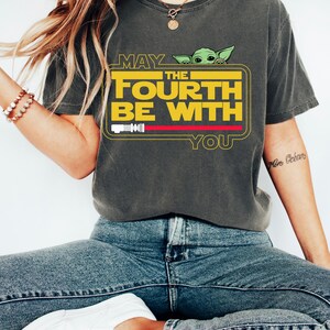 May include: Dark grey t-shirt with the text "MAY THE FOURTH BE WITH YOU" in yellow, a green alien character, and a red lightsaber graphic. The shirt is worn with blue jeans and white shoes.