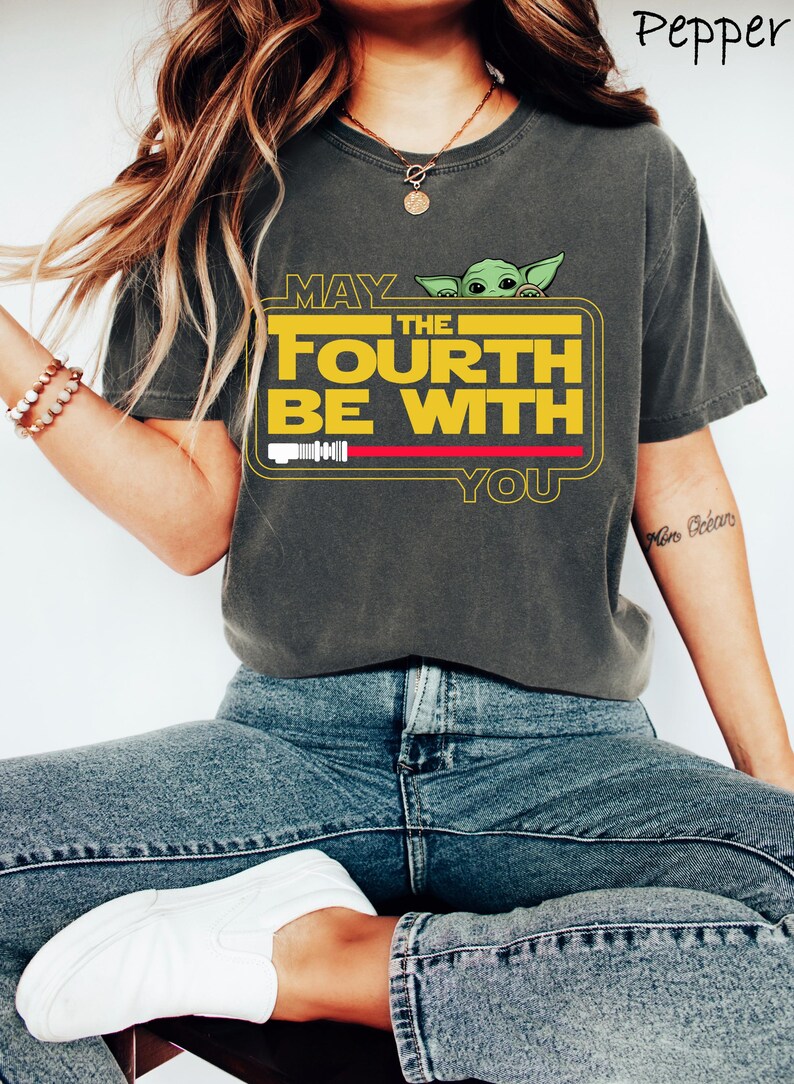 May include: Dark grey t-shirt with the text "MAY THE FOURTH BE WITH YOU" in yellow, a green alien character, and a red lightsaber graphic. The shirt is worn with blue jeans and white shoes.