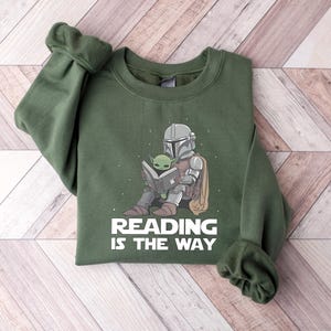 May include: Green sweatshirt featuring a graphic of The Mandalorian and Grogu reading a book. The text "READING IS THE WAY" is printed below the illustration. The sweatshirt has long sleeves and a crew neck.
