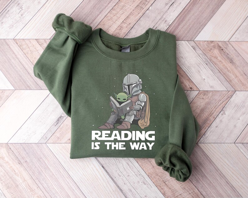 May include: Green sweatshirt featuring a graphic of The Mandalorian and Grogu reading a book. The text "READING IS THE WAY" is printed below the illustration. The sweatshirt has long sleeves and a crew neck.
