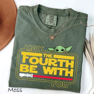 May include: A moss green t-shirt with a yellow and red graphic that reads "MAY THE FOURTH BE WITH YOU", featuring a cartoon character. A gold necklace and a brown hat are also visible in the image.
