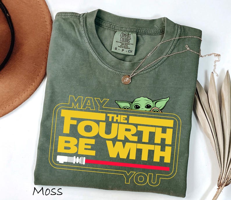 May include: A moss green t-shirt with a yellow and red graphic that reads "MAY THE FOURTH BE WITH YOU", featuring a cartoon character. A gold necklace and a brown hat are also visible in the image.