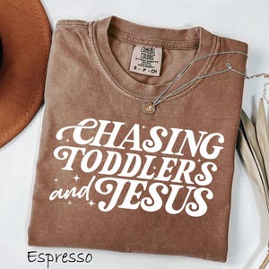 May include: Espresso-colored t-shirt with the text "CHASING TODDLERS and JESUS" in white, retro-style lettering. A silver necklace with a small pendant is draped across the shirt. The shirt is folded on a white surface.