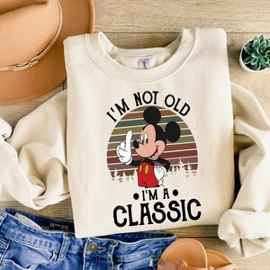 Puede incluir: Sudadera de color crema con un gr&aacute;fico de Mickey Mouse y el texto "I'M NOT OLD I'M A CLASSIC". El dise&ntilde;o incluye una puesta de sol retro y &aacute;rboles. La sudadera se combina con pantalones cortos de mezclilla azules y un sombrero beige.