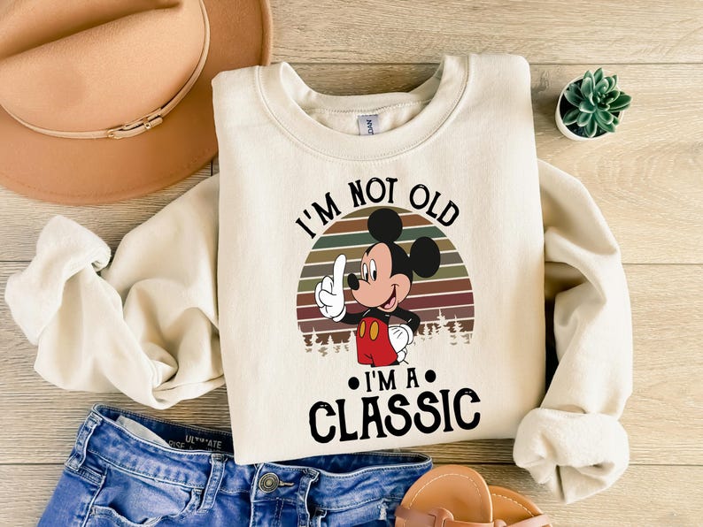 Puede incluir: Sudadera de color crema con un gr&aacute;fico de Mickey Mouse y el texto "I'M NOT OLD I'M A CLASSIC". El dise&ntilde;o incluye una puesta de sol retro y &aacute;rboles. La sudadera se combina con pantalones cortos de mezclilla azules y un sombrero beige.