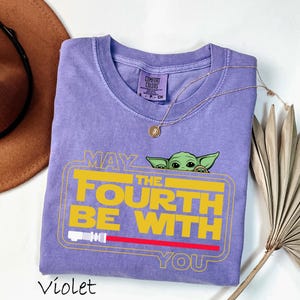 May include: A violet Comfort Colors t-shirt with a Baby Yoda graphic and the text "May the Fourth Be With You" in yellow and red. The shirt has a crew neck and short sleeves. A gold necklace is draped across the shirt.