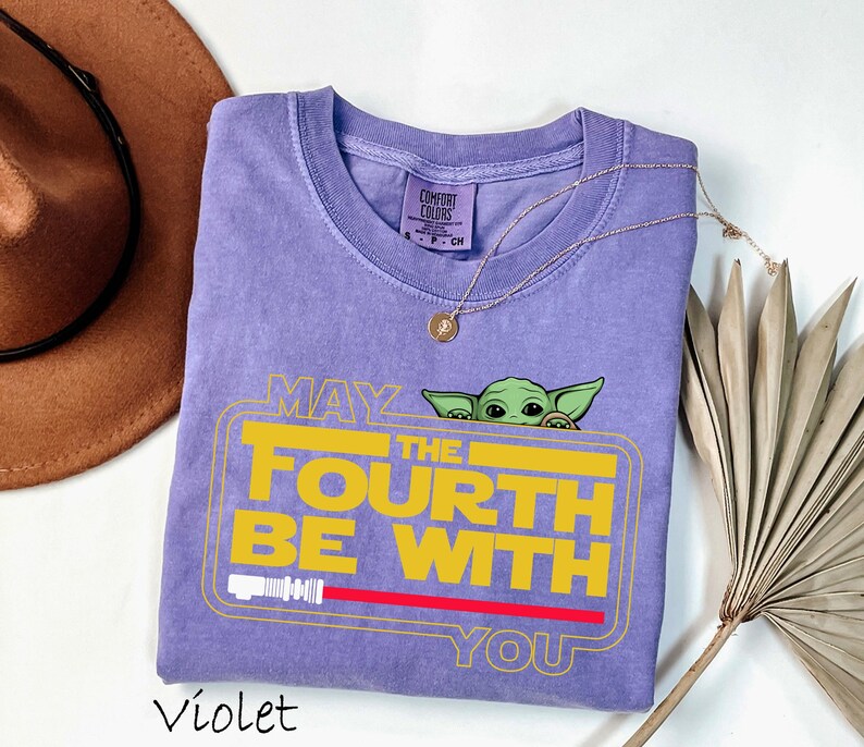 May include: A violet Comfort Colors t-shirt with a Baby Yoda graphic and the text "May the Fourth Be With You" in yellow and red. The shirt has a crew neck and short sleeves. A gold necklace is draped across the shirt.