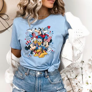 Może przedstawiać: Jasnoniebieski t-shirt z nadrukiem graficznym przedstawiającym Myszkę Miki, Myszkę Minnie, Kaczora Donalda, Goofy'ego i Pluto w patriotycznych czerwono-biało-niebieskich akcesoriach. Wz&oacute;r zawiera fajerwerki oraz imiona postaci napisane kursywą.