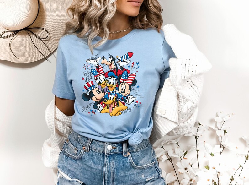 Może przedstawiać: Jasnoniebieski t-shirt z nadrukiem graficznym przedstawiającym Myszkę Miki, Myszkę Minnie, Kaczora Donalda, Goofy'ego i Pluto w patriotycznych czerwono-biało-niebieskich akcesoriach. Wz&oacute;r zawiera fajerwerki oraz imiona postaci napisane kursywą.