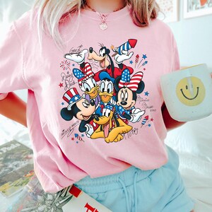 Może przedstawiać: Jasnor&oacute;żowy t-shirt z nadrukiem graficznym przedstawiającym postacie Disneya, w tym Myszkę Miki, Myszkę Minnie, Kaczora Donalda, Daisy, Goofy'ego i Pluto, noszące patriotyczne czerwono-biało-niebieskie akcesoria, fajerwerki oraz imiona.