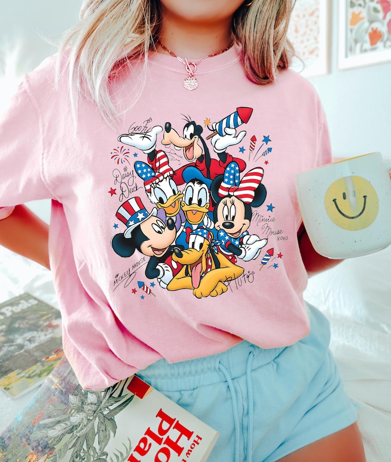 Może przedstawiać: Jasnor&oacute;żowy t-shirt z nadrukiem graficznym przedstawiającym postacie Disneya, w tym Myszkę Miki, Myszkę Minnie, Kaczora Donalda, Daisy, Goofy'ego i Pluto, noszące patriotyczne czerwono-biało-niebieskie akcesoria, fajerwerki oraz imiona.