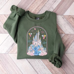 K&ouml;nnte beinhalten: Ein olivgr&uuml;nes Sweatshirt mit einer farbenfrohen Grafik eines Schlosses, Feuerwerks und einer blauen Cartoonfigur. Die Grafik wird von einem Bogen mit rosa und gelben Akzenten umrahmt. Das Sweatshirt hat lange &Auml;rmel.