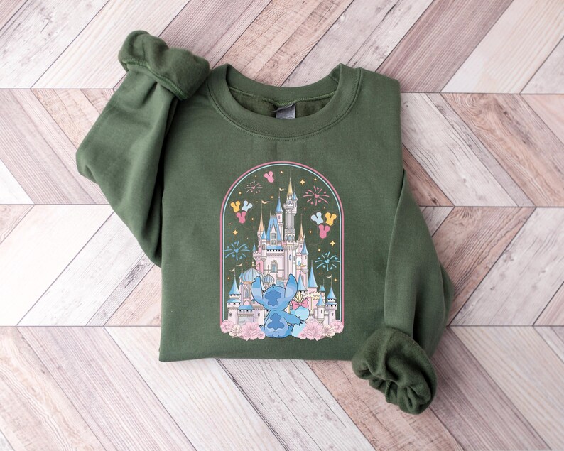 K&ouml;nnte beinhalten: Ein olivgr&uuml;nes Sweatshirt mit einer farbenfrohen Grafik eines Schlosses, Feuerwerks und einer blauen Cartoonfigur. Die Grafik wird von einem Bogen mit rosa und gelben Akzenten umrahmt. Das Sweatshirt hat lange &Auml;rmel.