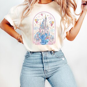 K&ouml;nnte beinhalten: Cremefarbenes T-Shirt mit einer Cartoon-Illustration eines blauen Charakters vor einem M&auml;rchenschloss. Das Design umfasst rosa Blumen, Feuerwerk und Ballons, alles in einem bogenf&ouml;rmigen Rahmen. Das Shirt wird mit hellblauen Jeans getragen.