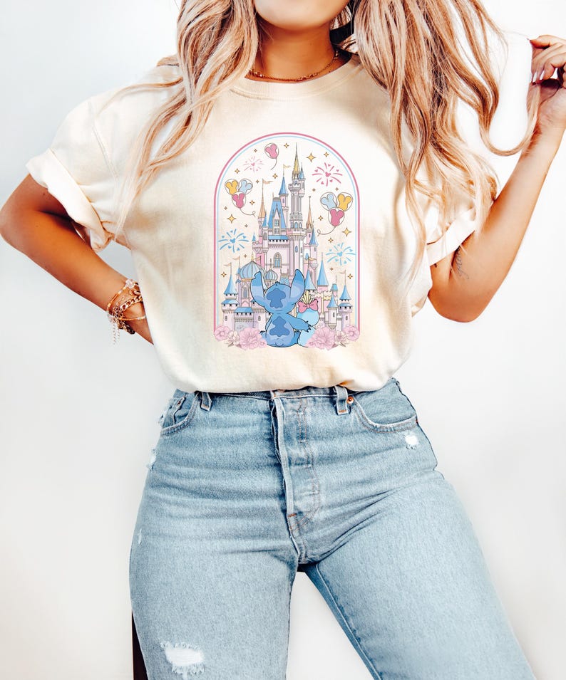 K&ouml;nnte beinhalten: Cremefarbenes T-Shirt mit einer Cartoon-Illustration eines blauen Charakters vor einem M&auml;rchenschloss. Das Design umfasst rosa Blumen, Feuerwerk und Ballons, alles in einem bogenf&ouml;rmigen Rahmen. Das Shirt wird mit hellblauen Jeans getragen.
