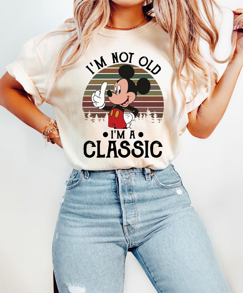 Puede incluir: Camiseta color crema con Mickey Mouse y el texto "I'M NOT OLD I'M A CLASSIC". Mickey Mouse levanta el dedo, sobre un dise&ntilde;o de puesta de sol. La camiseta se combina con vaqueros azul claro.