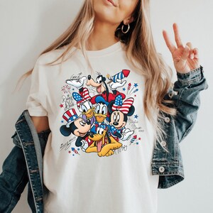 Może przedstawiać: Kremowy t-shirt z nadrukiem graficznym przedstawiającym postacie Disneya, w tym Myszkę Miki, Myszkę Minnie, Kaczora Donalda, Daisy, Goofy'ego i Pluto. Postacie są ozdobione patriotycznymi elementami w kolorach czerwonym, białym i niebieskim.