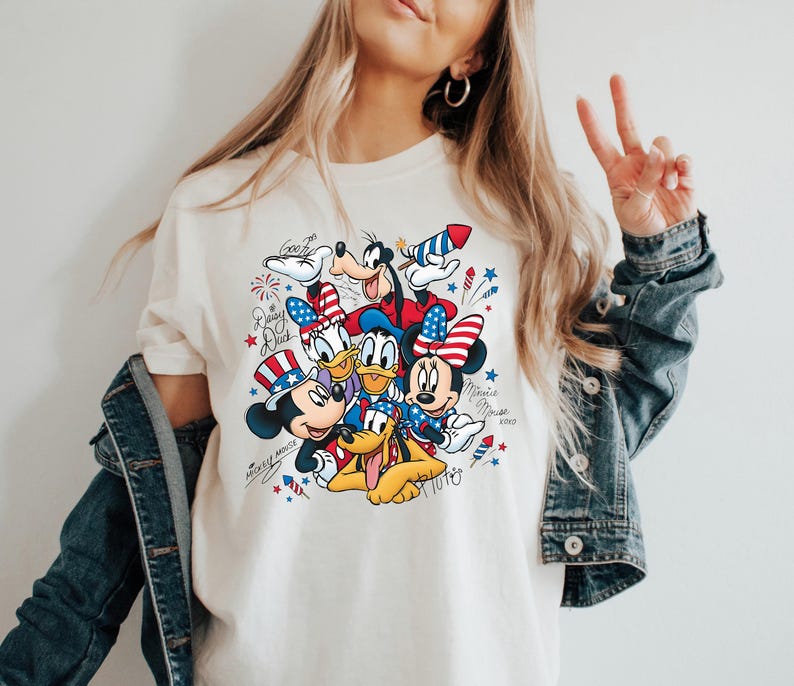 Może przedstawiać: Kremowy t-shirt z nadrukiem graficznym przedstawiającym postacie Disneya, w tym Myszkę Miki, Myszkę Minnie, Kaczora Donalda, Daisy, Goofy'ego i Pluto. Postacie są ozdobione patriotycznymi elementami w kolorach czerwonym, białym i niebieskim.