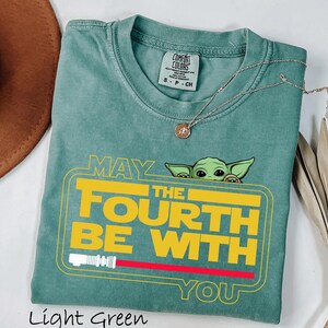 May include: Light green t-shirt with the text "MAY THE FOURTH BE WITH YOU" in yellow and white, featuring a cartoon character. A red lightsaber graphic is below the text. A silver necklace with a pendant is on the shirt.