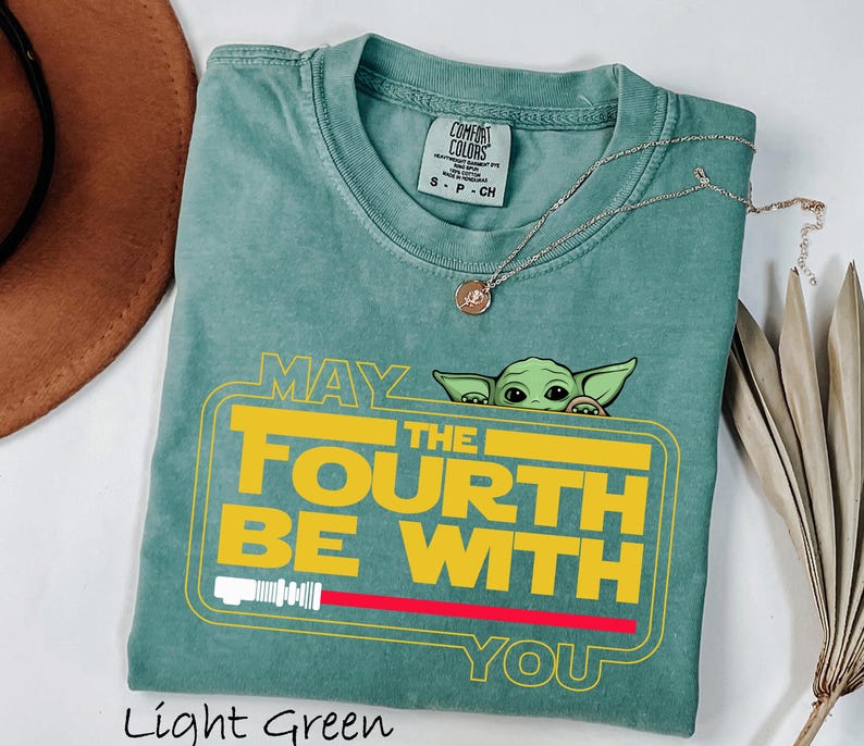 May include: Light green t-shirt with the text "MAY THE FOURTH BE WITH YOU" in yellow and white, featuring a cartoon character. A red lightsaber graphic is below the text. A silver necklace with a pendant is on the shirt.