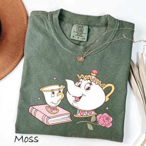 May include: Moss green t-shirt featuring a cartoon illustration of a teapot and teacup with a pink rose. The shirt has a crew neck and short sleeves. The word "Moss" is printed at the bottom of the shirt.