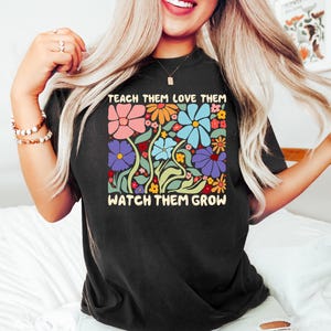 May include: Black t-shirt with a colorful floral design featuring pink, orange, blue, and purple flowers. The text "TEACH THEM LOVE THEM WATCH THEM GROW" is printed above and below the floral graphic.