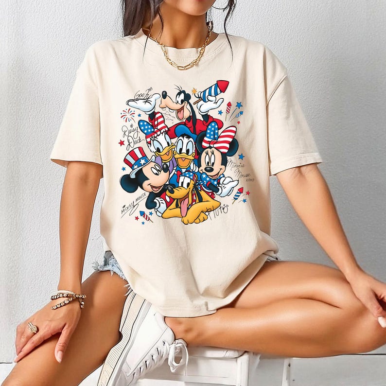 Może przedstawiać: Beżowy t-shirt z kr&oacute;tkim rękawem z nadrukiem graficznym przedstawiającym postacie Disneya, w tym Myszkę Miki, Myszkę Minnie, Kaczora Donalda, Daisy, Goofy'ego i Pluto. Postacie mają na sobie patriotyczne akcesoria w kolorach czerwonym, białym i niebieskim.