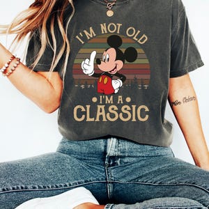 Puede incluir: Camiseta gris oscuro con un dise&ntilde;o de Mickey Mouse y el texto "I'M NOT OLD I'M A CLASSIC". El dise&ntilde;o muestra a Mickey Mouse se&ntilde;alando hacia arriba sobre un fondo de puesta de sol retro. Una camiseta informal.