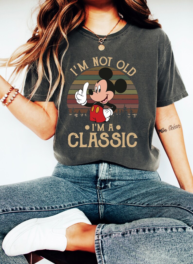 Puede incluir: Camiseta gris oscuro con un dise&ntilde;o de Mickey Mouse y el texto "I'M NOT OLD I'M A CLASSIC". El dise&ntilde;o muestra a Mickey Mouse se&ntilde;alando hacia arriba sobre un fondo de puesta de sol retro. Una camiseta informal.