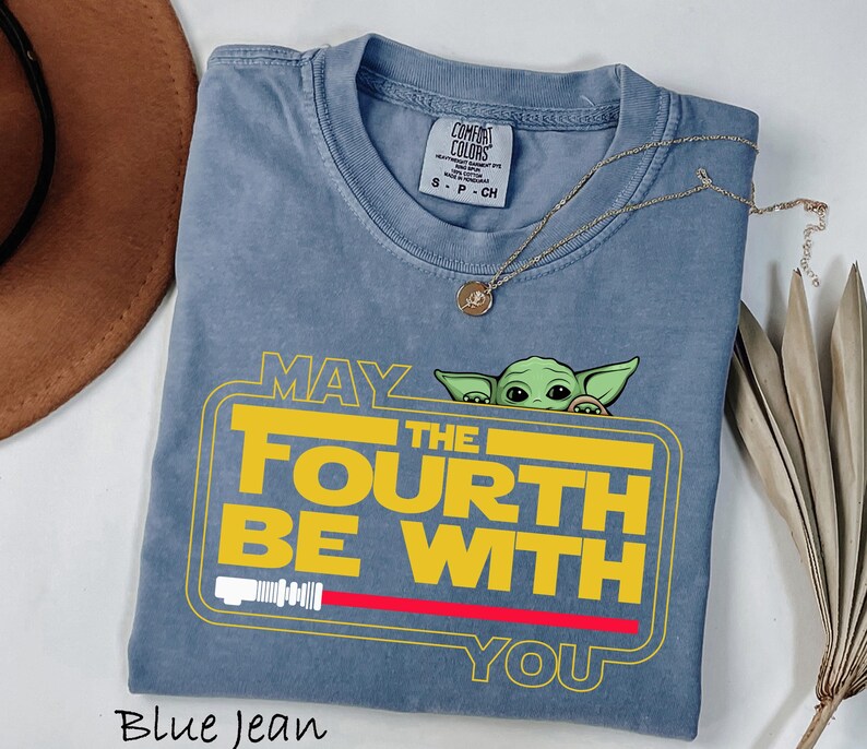 May include: Blue jean coloured t-shirt with the text "MAY THE FOURTH BE WITH YOU" in yellow and white, featuring a green cartoon character. A red lightsaber graphic is below the text. A gold necklace is draped across the shirt.