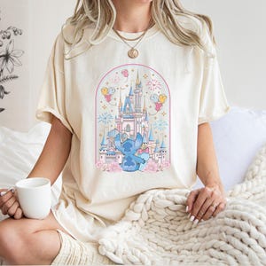 K&ouml;nnte beinhalten: Cremefarbenes T-Shirt mit einer Cartoon-Illustration eines blauen Charakters vor einem M&auml;rchenschloss. Das Design umfasst rosa und blaue Akzente, Feuerwerk und Luftballons. Das Shirt besteht aus einem weichen Material.