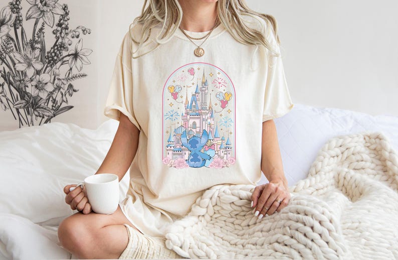 K&ouml;nnte beinhalten: Cremefarbenes T-Shirt mit einer Cartoon-Illustration eines blauen Charakters vor einem M&auml;rchenschloss. Das Design umfasst rosa und blaue Akzente, Feuerwerk und Luftballons. Das Shirt besteht aus einem weichen Material.