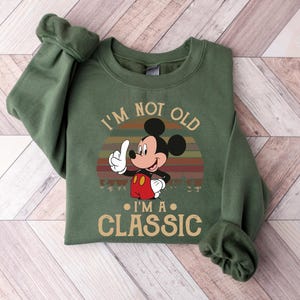 Puede incluir: Sudadera verde con Mickey Mouse y el texto "I'm not old, I'm a classic". El dise&ntilde;o incluye un atardecer retro. Mickey Mouse hace una se&ntilde;al de pulgar hacia arriba.