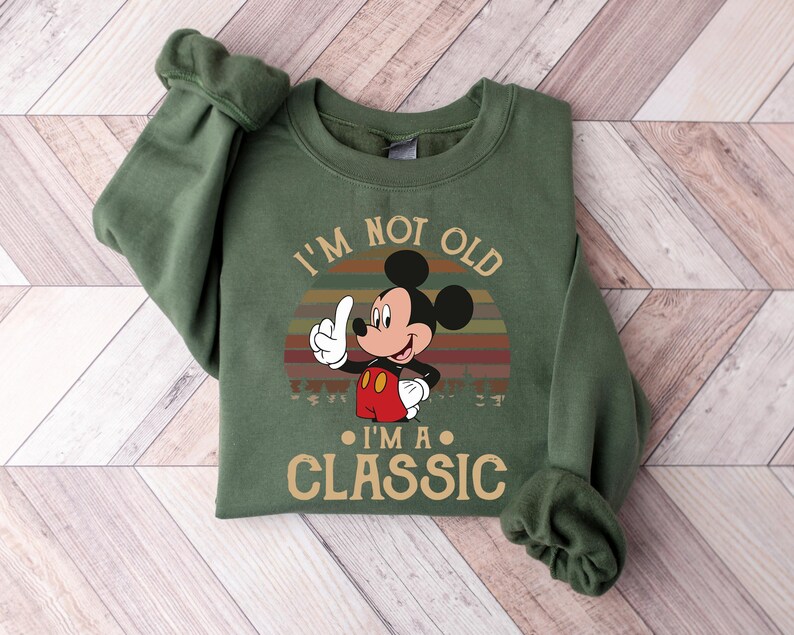Puede incluir: Sudadera verde con Mickey Mouse y el texto "I'm not old, I'm a classic". El dise&ntilde;o incluye un atardecer retro. Mickey Mouse hace una se&ntilde;al de pulgar hacia arriba.