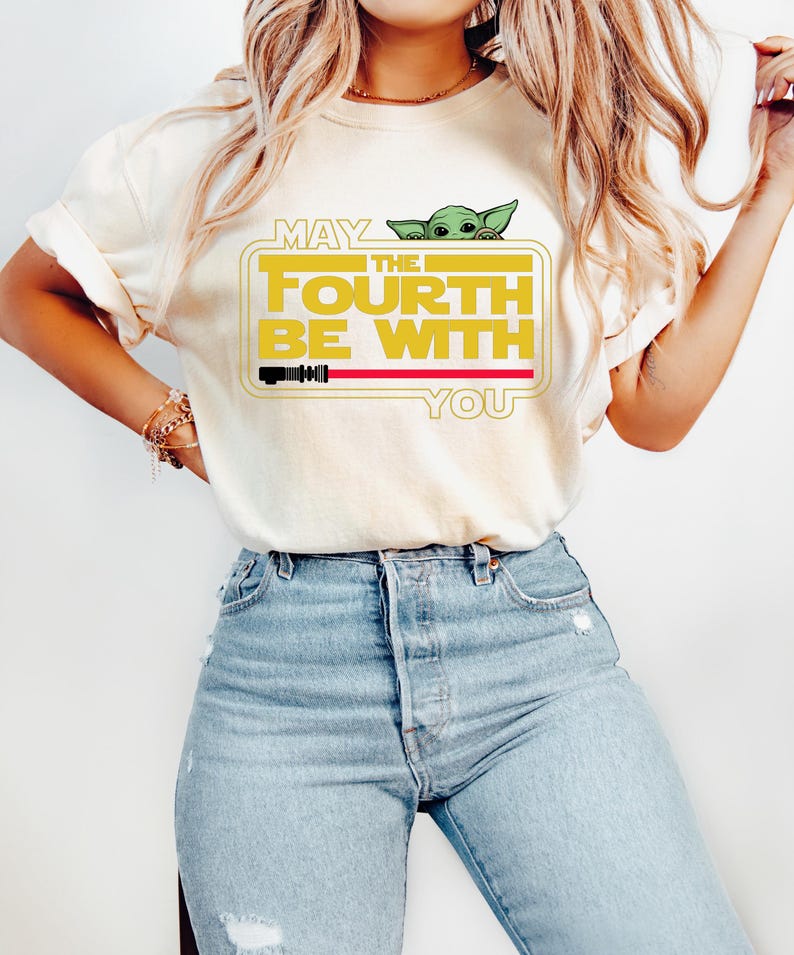 May include: Cream-coloured t-shirt with the text "MAY THE FOURTH BE WITH YOU" in yellow and a green cartoon character. The design includes a red lightsaber and a yellow rectangular frame. The shirt is paired with light blue jeans.