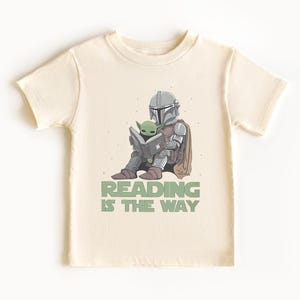 May include: Cream-colored t-shirt featuring a cartoon illustration of a Mandalorian character and Grogu reading a book. The text "READING IS THE WAY" is printed below the characters in green. The shirt has short sleeves and a crew neck.