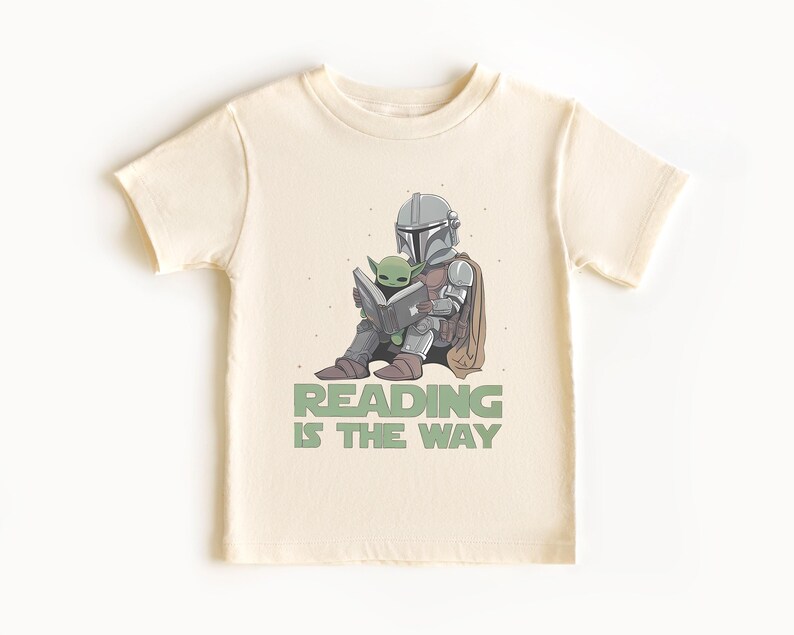 May include: Cream-colored t-shirt featuring a cartoon illustration of a Mandalorian character and Grogu reading a book. The text "READING IS THE WAY" is printed below the characters in green. The shirt has short sleeves and a crew neck.