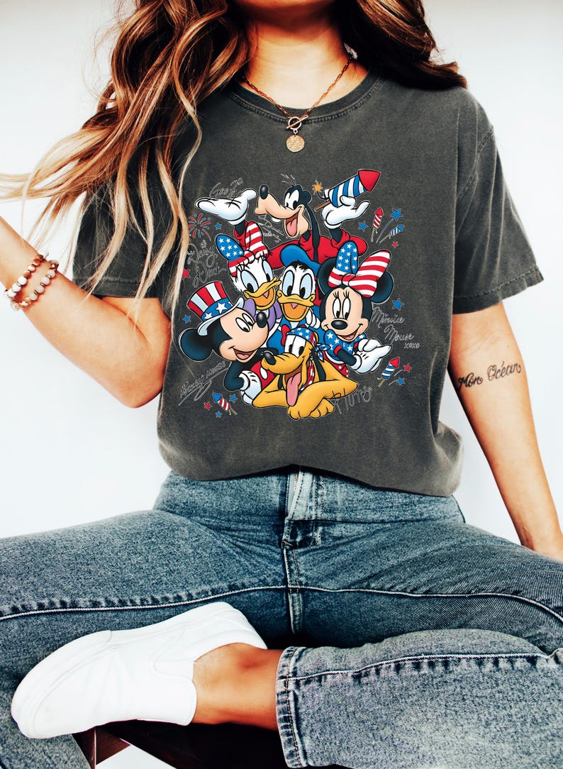 Może przedstawiać: Ciemnoszary t-shirt z kr&oacute;tkim rękawem z nadrukiem graficznym przedstawiającym Myszkę Miki, Myszkę Minnie, Kaczora Donalda, Daisy, Goofy'ego i Pluto w patriotycznych czerwono-biało-niebieskich akcesoriach, w otoczeniu ilustracji fajerwerk&oacute;w i gwiazd.