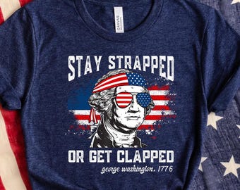 Puede incluir: Una camiseta azul marino con un gráfico de George Washington con gafas de sol y una bandana con la bandera estadounidense. El texto "Stay Strapped or Get Clapped George Washington, 1776" está impreso en la camiseta.
