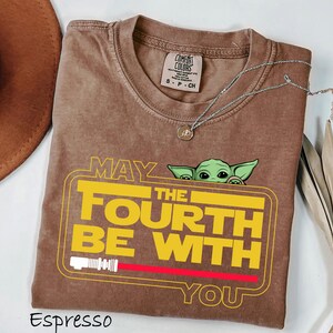 May include: Espresso-coloured t-shirt with a yellow and red graphic that reads "MAY THE FOURTH BE WITH YOU" and a green cartoon character. A silver necklace with a pendant is draped across the shirt.