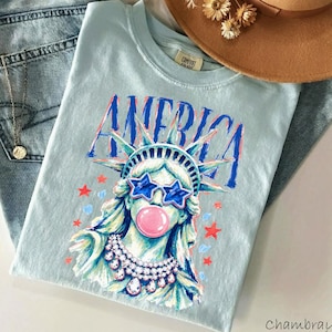 May include: A light blue t-shirt featuring a graphic of the Statue of Liberty wearing star-shaped blue sunglasses and blowing a pink bubble. The word AMERICA is printed in blue above the statue, which is adorned with a white beaded necklace.