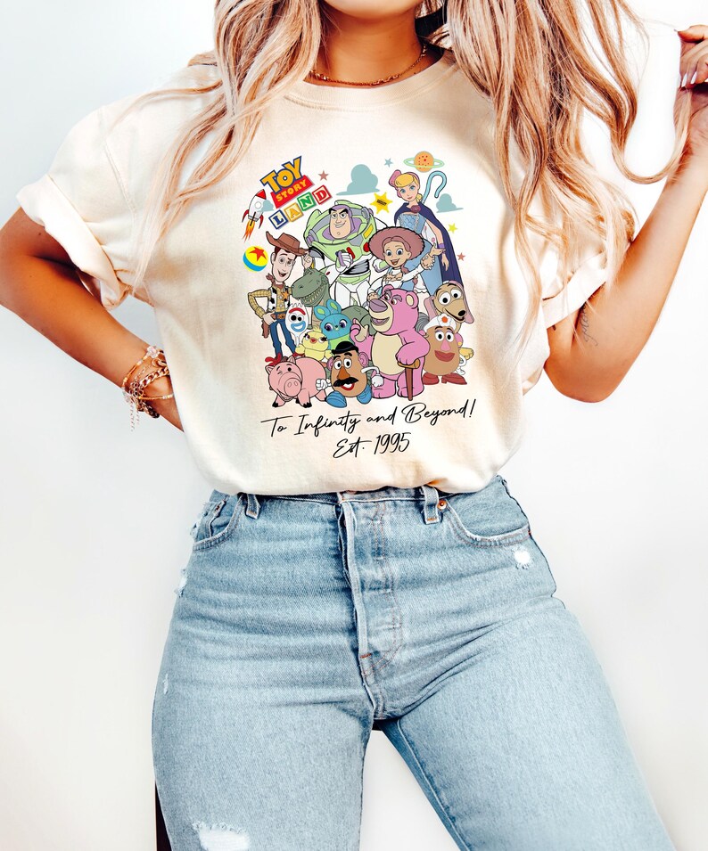 May include: Cream-colored t-shirt featuring a colorful graphic of Toy Story characters. The design includes Woody, Buzz Lightyear, and other characters from the movie, with the text "Toy Story Land" and "To Infinity and Beyond! Est. 1995".